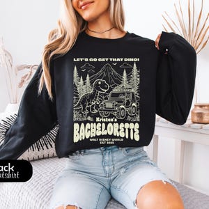 Op de afbeelding: Een zwart sweatshirt met lange mouwen en een cr&egrave;mekleurige grafische print. Het ontwerp toont een dinosaurus, een jeep, dennenbomen en een berg, met de tekst: LET'S GO GET THAT DINO! Kristen's BACHELORETTE WALT DISNEY WORLD EST 2025.