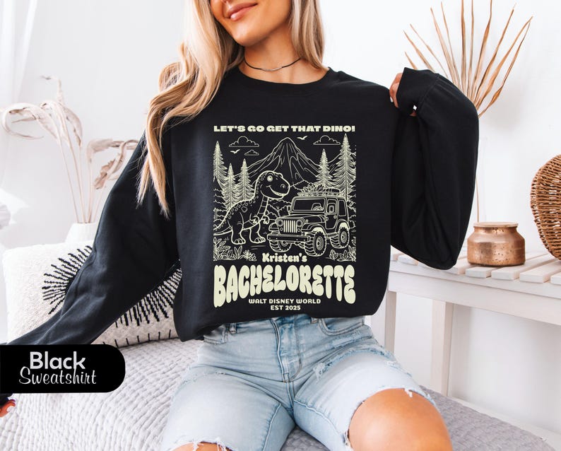 Op de afbeelding: Een zwart sweatshirt met lange mouwen en een cr&egrave;mekleurige grafische print. Het ontwerp toont een dinosaurus, een jeep, dennenbomen en een berg, met de tekst: LET'S GO GET THAT DINO! Kristen's BACHELORETTE WALT DISNEY WORLD EST 2025.