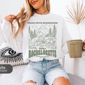 Op de afbeelding: Een wit sweatshirt met lange mouwen en een groene grafische print van een dinosaurus, een jeep, bergen en bomen. De tekst luidt ROAR INTO MARRIAGE, Smily's BACHELORETTE, COWICHAN VALLEY EST 2025 en staat centraal op de voorzijde van het kledingstuk.
