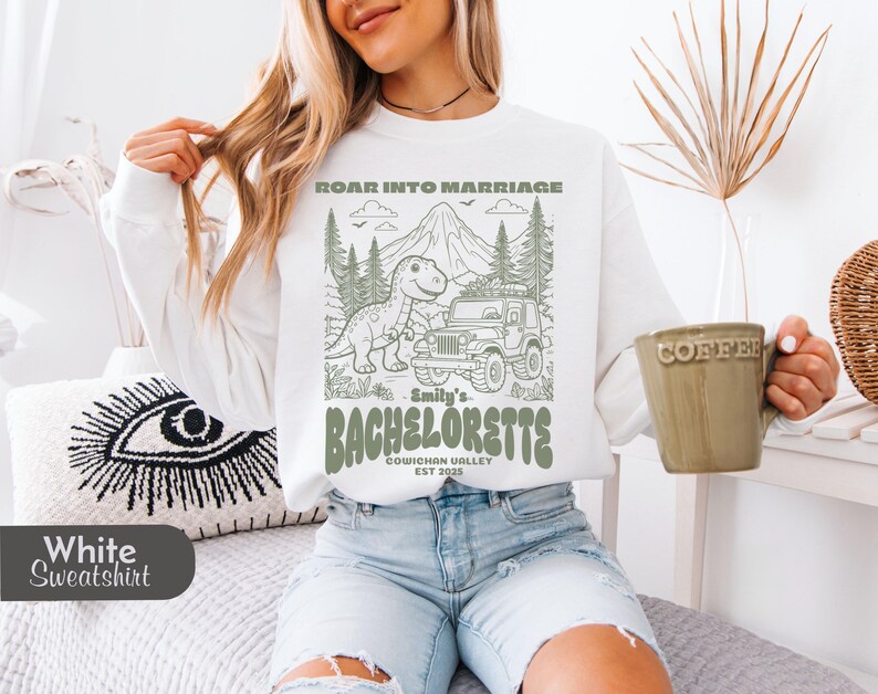 Op de afbeelding: Een wit sweatshirt met lange mouwen en een groene grafische print van een dinosaurus, een jeep, bergen en bomen. De tekst luidt ROAR INTO MARRIAGE, Smily's BACHELORETTE, COWICHAN VALLEY EST 2025 en staat centraal op de voorzijde van het kledingstuk.