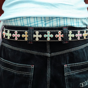 Könnte beinhalten: Schwarzer Denim-Gürtel mit bunten Kreuzverzierungen. Die Kreuze sind mit verschiedenfarbigen Strasssteinen verziert, darunter rosa, blau, grün und gelb. Der Gürtel wird mit dunkelblauen Jeans und einem hellblauen Hemd getragen.