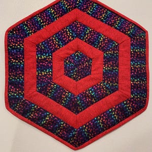 May include: A hexagonal table runner with a red and black color scheme. The runner features a repeating pattern of rainbow musical notes on a black background. The runner has a red border and a red center.