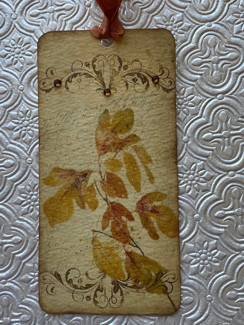 May include: A vintage-style bookmark featuring a pressed leaf design. The bookmark is decorated with a brown ribbon and has a distressed, aged look. The background features handwritten script.