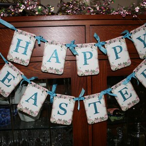 May include: A banner with the word "Happy Easter" in blue letters on a white background with floral designs. The banner is strung with blue ribbon and hung on a wooden cabinet.