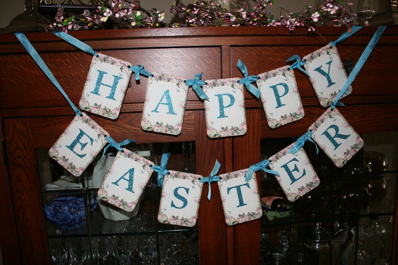 May include: A banner with the word "Happy Easter" in blue letters on a white background with floral designs. The banner is strung with blue ribbon and hung on a wooden cabinet.