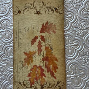 May include: A vintage-style bookmark with a brown background and a decorative border. The bookmark features a design of autumn leaves in shades of orange, red, and yellow. The leaves are printed on a white background and have a faded, antique look. The bookmark is accented with small, brown rhinestones.