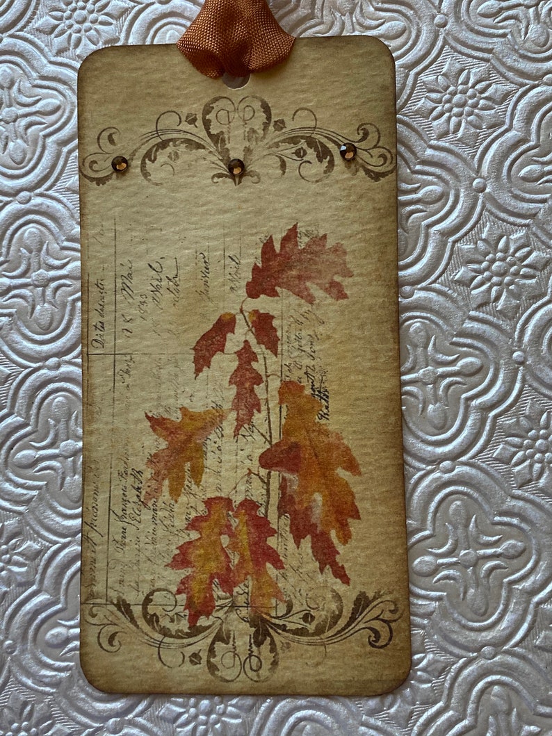 May include: A vintage-style bookmark with a brown background and a decorative border. The bookmark features a design of autumn leaves in shades of orange, red, and yellow. The leaves are printed on a white background and have a faded, antique look. The bookmark is accented with small, brown rhinestones.
