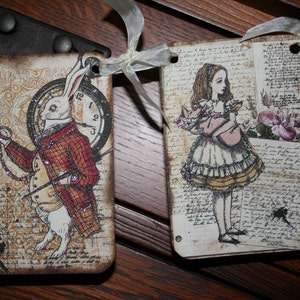 May include: Two decorative paper banners with a vintage aesthetic. One features a white rabbit wearing a red and black plaid vest and holding a pocket watch. The other features a girl in a white dress with a pink bodice and a crown of flowers.