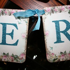 May include: A banner with the letters "ER" in turquoise glitter on a white background. The banner is decorated with a floral pattern and blue birds.