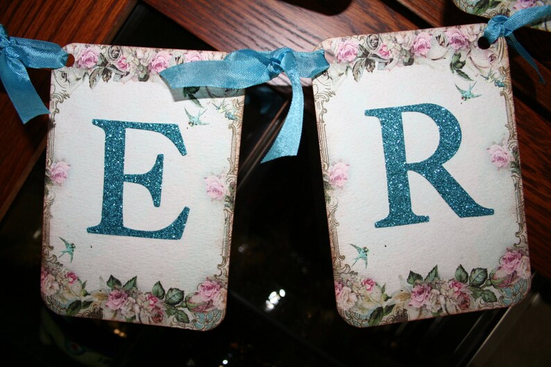 May include: A banner with the letters "ER" in turquoise glitter on a white background. The banner is decorated with a floral pattern and blue birds.