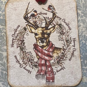 May include: A rectangular Christmas ornament featuring a deer with a red and white plaid scarf, surrounded by a wreath and two birds. The words "Merry Christmas" are printed around the edge. A red ribbon is attached for hanging.