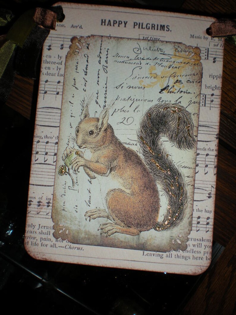 Pode incluir: Uma etiqueta retangular com um design vintage com um esquilo marrom segurando uma noz. A etiqueta tem um aspecto envelhecido com escrita e notas musicais no fundo. O texto "HAPPY PILGRIMS" est&aacute; no topo, com uma fita marrom e verde.