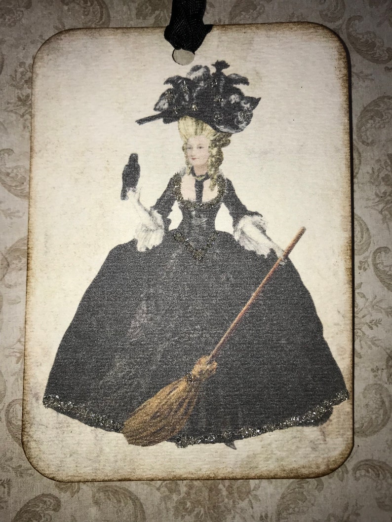 May include: A rectangular wooden ornament featuring a vintage illustration of a woman in a black gown and elaborate hat, holding a broom and a bird. The image has a distressed, aged appearance, with a black ribbon for hanging.