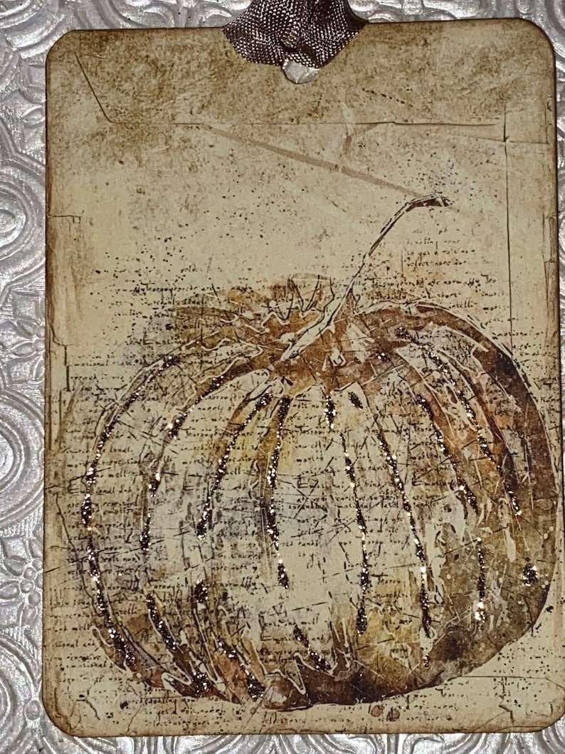 Op de afbeelding: Een rechthoekig label met een vintage pompoenillustratie. De pompoen is bruin en goud met glitteraccenten, tegen een achtergrond van verouderd papier met schrift. Bovenop is een bruin lint bevestigd.