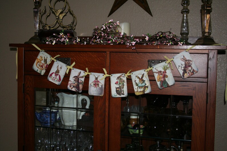 May include: A vintage-style Easter bunny banner with yellow ribbon and a wooden cabinet in the background. The banner features illustrations of bunnies in various poses, including holding carrots and wearing hats.