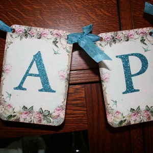 May include: A blue and white banner with the letters "A" and "P" in a sparkly blue font. The banner is decorated with floral designs and small blue birds.