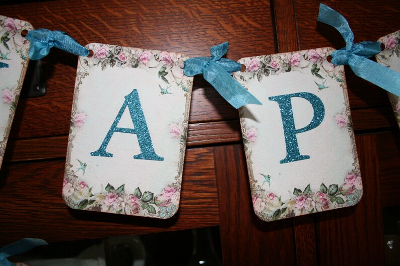 May include: A blue and white banner with the letters "A" and "P" in a sparkly blue font. The banner is decorated with floral designs and small blue birds.