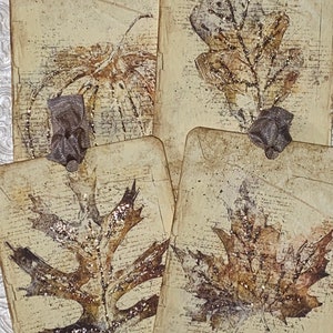 Op de afbeelding: Vier handgemaakte papieren labels met bladvormgeving. De labels zijn beige met een vintage script achtergrond en bruine blad illustraties. Elk label heeft een klein, decoratief zilveren lint. De bladeren hebben glitter accenten.