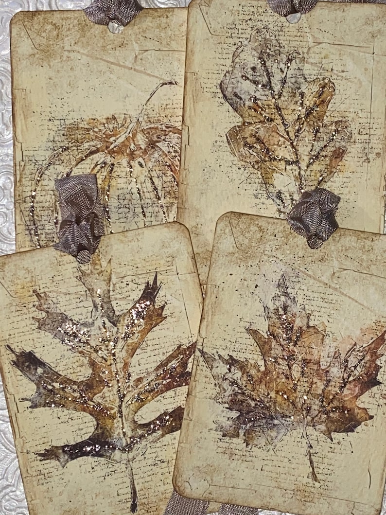 Op de afbeelding: Vier handgemaakte papieren labels met bladvormgeving. De labels zijn beige met een vintage script achtergrond en bruine blad illustraties. Elk label heeft een klein, decoratief zilveren lint. De bladeren hebben glitter accenten.