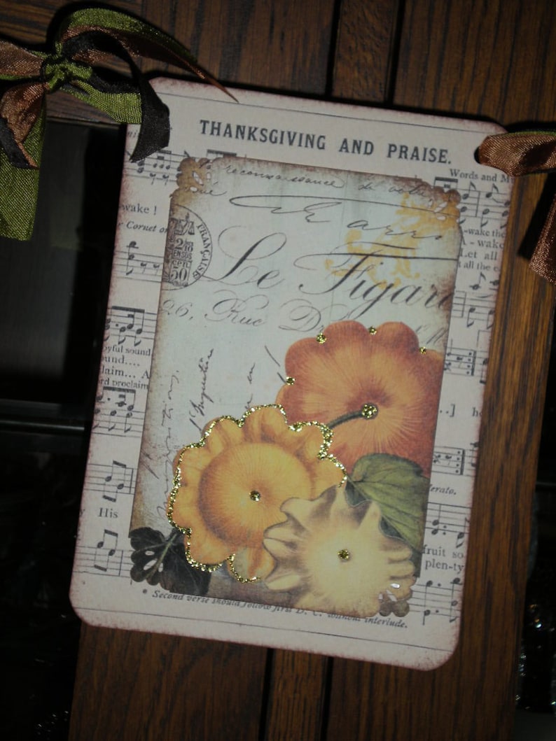 Pode incluir: Uma etiqueta retangular com tema de A&ccedil;&atilde;o de Gra&ccedil;as com as palavras "THANKSGIVING AND PRAISE". Apresenta um design vintage com elementos florais e de notas musicais, e imagens de ab&oacute;boras. A etiqueta &eacute; adornada com glitter dourado e fita.