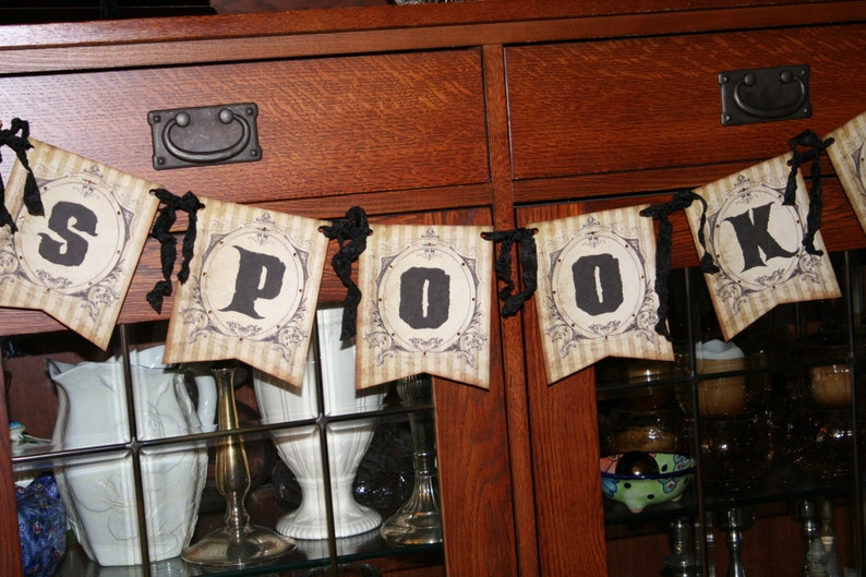 May include: A decorative banner featuring five individual pennants strung together with black ribbon. Each pennant has a beige striped background with a circular frame containing a single black letter, spelling out the word SPOOK in sequence.