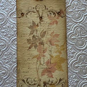 May include: A vintage-style tag with a brown background and a floral design. The tag has a brown ribbon attached to the top and features a floral design with brown and pink leaves. The tag has a handwritten inscription that reads "Autumn Leaves".