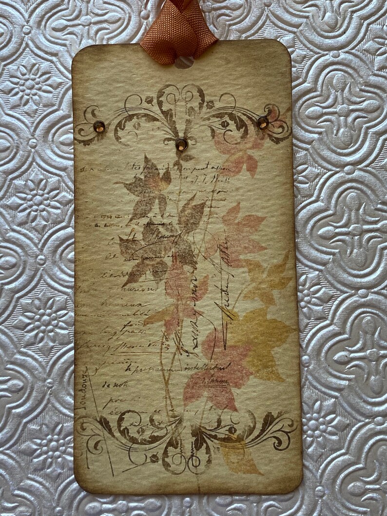 May include: A vintage-style tag with a brown background and a floral design. The tag has a brown ribbon attached to the top and features a floral design with brown and pink leaves. The tag has a handwritten inscription that reads "Autumn Leaves".