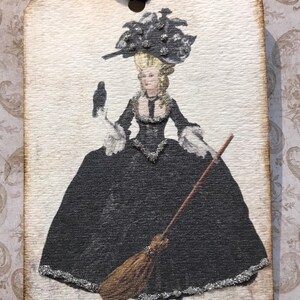 May include: A rectangular tag with a vintage illustration of a woman in a black gown, holding a raven and a broom. The woman wears a large hat and the tag has a black ribbon for hanging. The edges of the tag are brown.