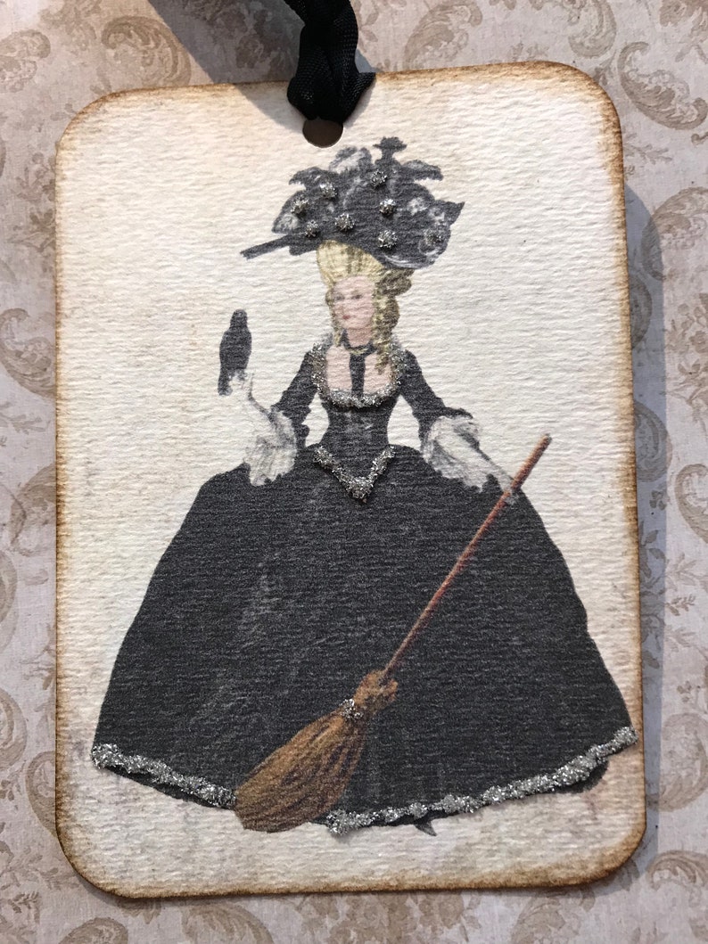 May include: A rectangular tag with a vintage illustration of a woman in a black gown, holding a raven and a broom. The woman wears a large hat and the tag has a black ribbon for hanging. The edges of the tag are brown.