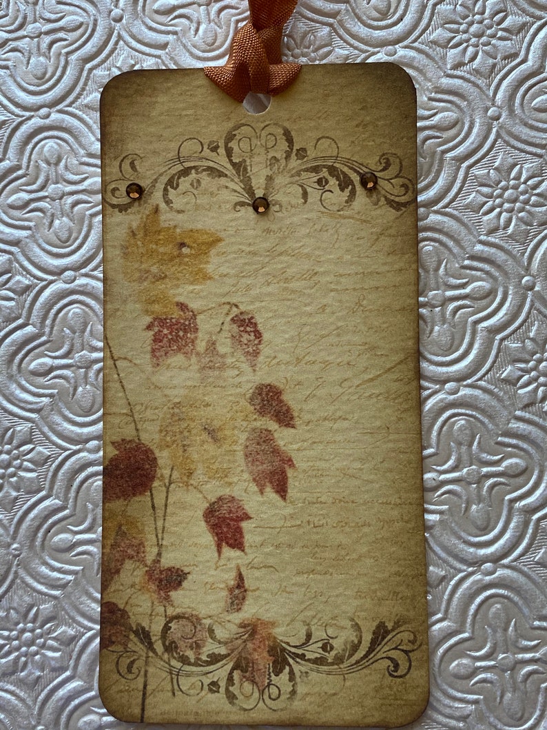 May include: A vintage-style gift tag with a brown background and a design of autumn leaves in shades of red, orange, and yellow. The tag has a decorative border and a hole at the top for hanging.