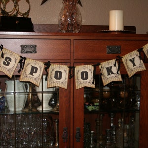 May include: A decorative banner featuring eight individual pennants strung together with black ribbon. Each pennant displays a vintage-style beige and brown striped background with black text spelling out S-P-O-O-K-Y, flanked by two skull illustrations.