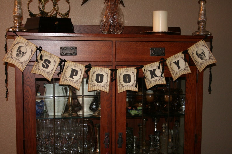May include: A decorative banner featuring eight individual pennants strung together with black ribbon. Each pennant displays a vintage-style beige and brown striped background with black text spelling out S-P-O-O-K-Y, flanked by two skull illustrations.