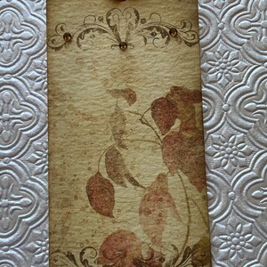 May include: A vintage-style tag with a brown background and a floral design. The tag has a brown ribbon attached to the top and features a faded image of leaves in shades of brown and red.