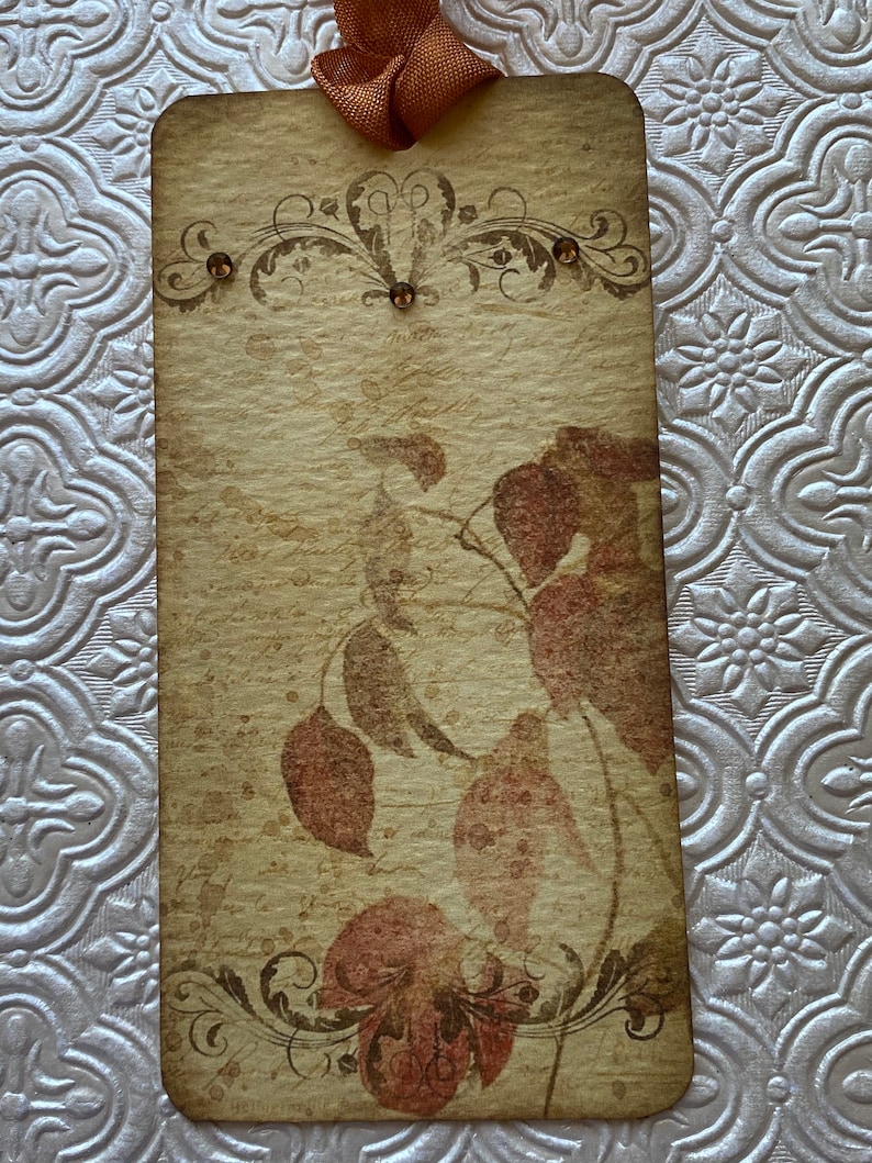 May include: A vintage-style tag with a brown background and a floral design. The tag has a brown ribbon attached to the top and features a faded image of leaves in shades of brown and red.