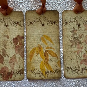 May include: Three vintage-style gift tags with brown ribbon and gold accents. Each tag features a different image of autumn leaves in shades of brown, yellow, and pink.