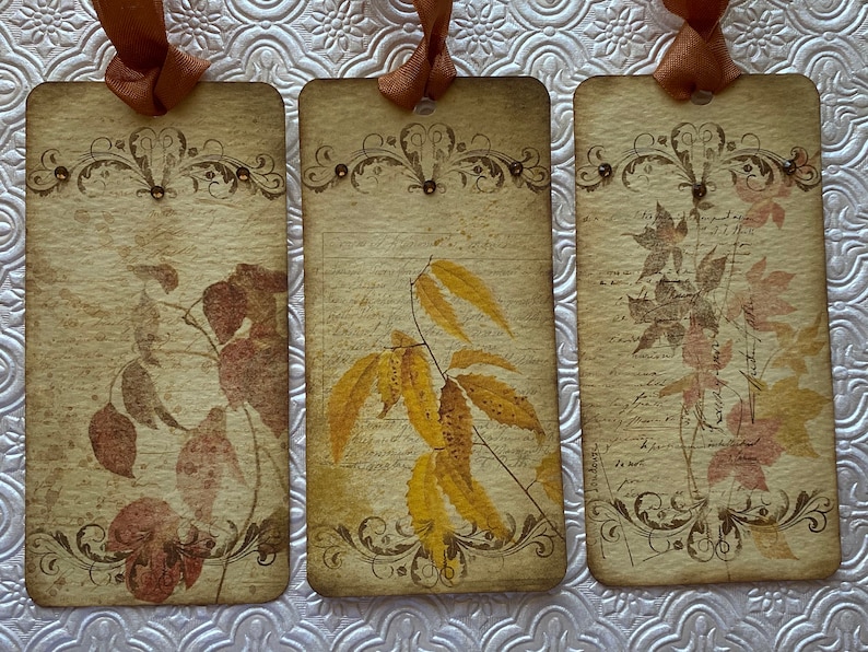 May include: Three vintage-style gift tags with brown ribbon and gold accents. Each tag features a different image of autumn leaves in shades of brown, yellow, and pink.