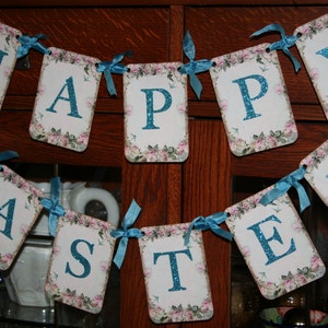May include: A banner with the words "Happy Easter" in blue glitter letters on a white background with pink and green floral designs. The banner is strung with blue ribbon.