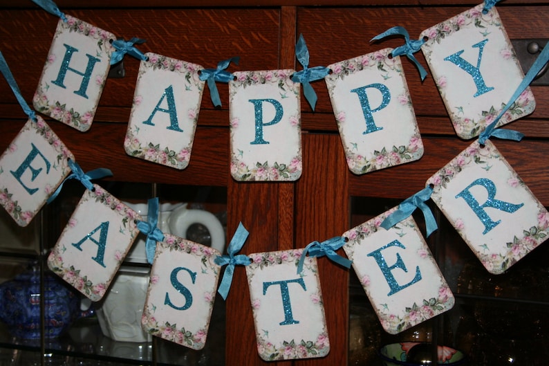 May include: A banner with the words "Happy Easter" in blue glitter letters on a white background with pink and green floral designs. The banner is strung with blue ribbon.