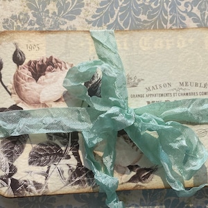 May include: A rectangular card with rounded corners, featuring a vintage rose design in sepia tones. The card is tied with a light blue ribbon. Text includes "1905" and French phrases.