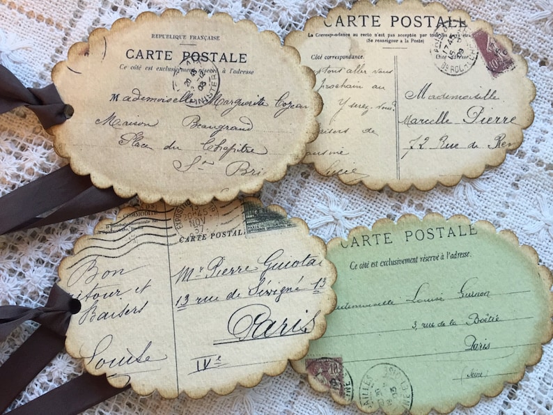 Puede incluir: Cuatro postales de estilo vintage con bordes festoneados y lazos de cinta marr&oacute;n. Las postales presentan escritura manuscrita, sellos y las palabras "CARTE POSTALE" en franc&eacute;s. Las postales son en tonos beige y verde.