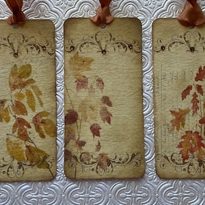 May include: Three vintage-style bookmarks with an autumn theme. Each bookmark features a different design of pressed autumn leaves on a cream-coloured background with a decorative border.