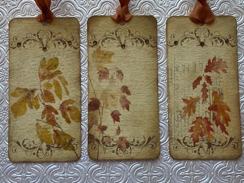 May include: Three vintage-style bookmarks with an autumn theme. Each bookmark features a different design of pressed autumn leaves on a cream-coloured background with a decorative border.
