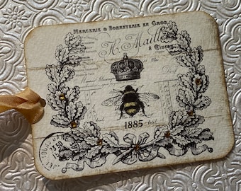 Op de afbeelding: Een vintage-stijl tag met een zwart-witte illustratie van een bij en eikenbladeren. De tag heeft de tekst "MERCERIS & BORMSTERIK EN GROS H. Maille à Ciura (M) 1885" erop gedrukt.