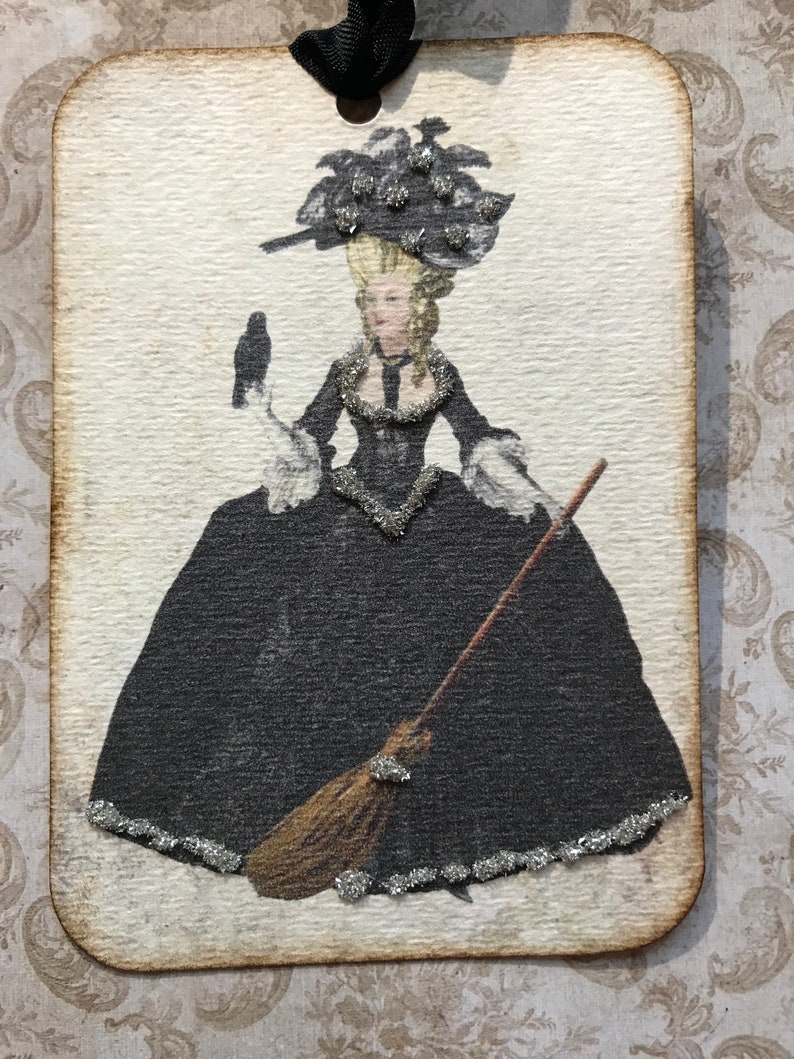 May include: A rectangular tag with a vintage illustration of a woman in a black gown, holding a broom and a bird. The image has silver glitter accents and a black ribbon for hanging. The tag has a distressed, aged appearance.