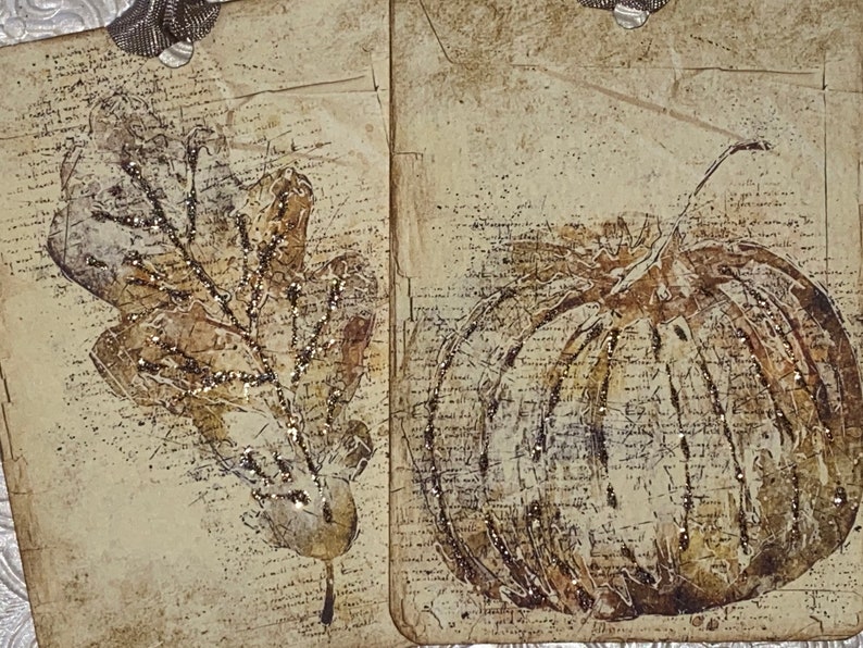 Op de afbeelding: Twee decoratieve labels met herfstthema's. E&eacute;n toont een gedetailleerd blad, de andere een pompoen. Beide zijn in bruin- en beigetinten, met een vintage, getextureerde uitstraling. Glitteraccenten voegen sprankeling toe. De labels zijn versierd met zilveren lint.