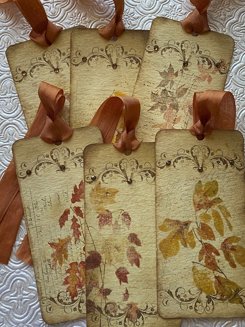 May include: Six vintage-style gift tags with brown ribbon. Each tag features a different design of autumn leaves in shades of brown, orange, and red. The tags are printed on cream-coloured paper with a distressed look.