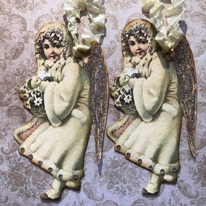 May include: Two vintage-style angel ornaments with glitter wings and white dresses. Each angel holds a bouquet of flowers and wears a white fur trim.