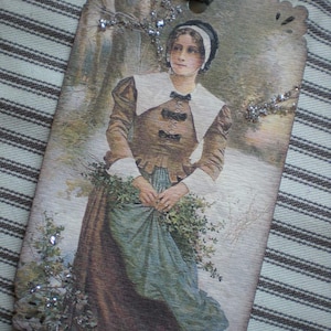 May include: A vintage-style tag featuring a woman in a historical dress, holding greenery in a snowy landscape. The tag has a decorative edge, embellished with glitter, and a gray ribbon for hanging. The woman is wearing a brown dress with a white collar.