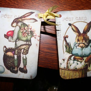 May include: Two vintage postcards featuring illustrations of rabbits. The first postcard shows a rabbit holding a red egg and a basket of eggs. The second postcard shows a rabbit wearing a blue apron and holding a basket of eggs. Both postcards have the text "POST CARD" printed on them.