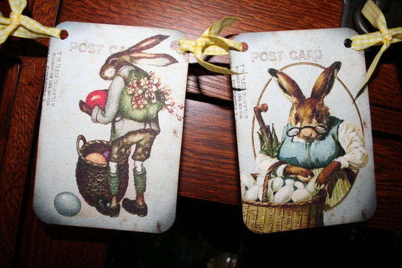 May include: Two vintage postcards featuring illustrations of rabbits. The first postcard shows a rabbit holding a red egg and a basket of eggs. The second postcard shows a rabbit wearing a blue apron and holding a basket of eggs. Both postcards have the text "POST CARD" printed on them.